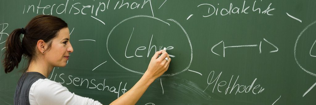 Eine Studierende der TU Chemnitz schreibt das Wort „Lehre“ an eine grüne Kreidetafel . Das Tafelbild rund um den zentralen Begriff Lehre zeigt weitere Verweise und Zusammenhänge mit den Begriffen interdisziplinär, Didaktik, Methoden und Wissenschaft.
