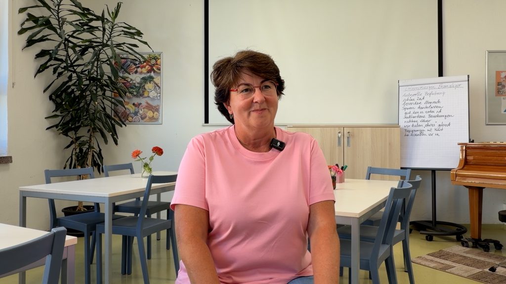 Eine Lehrerin sitzt in einem Klassenraum mit Tischen, Stühlen und Tafel und spricht über ihren Schulalltag.