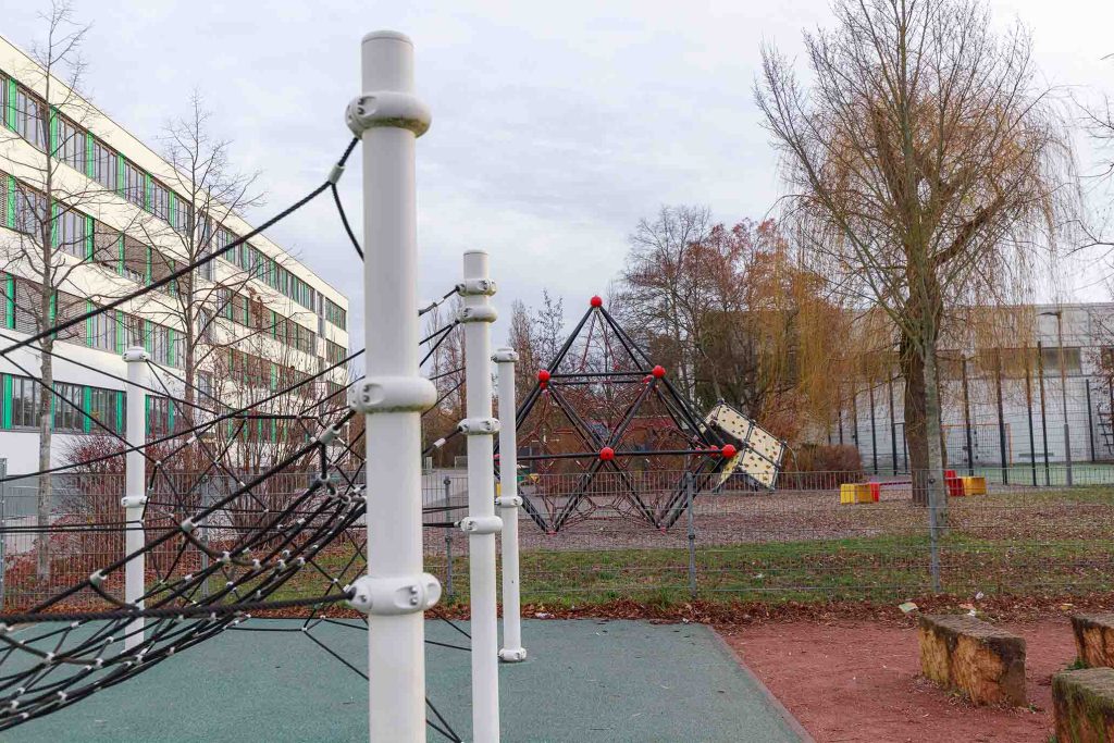 Ein Klettergerüst mit schwarzen Seilen und roten Knoten steht auf einem Schulhof neben einem mehrstöckigen Gebäude.